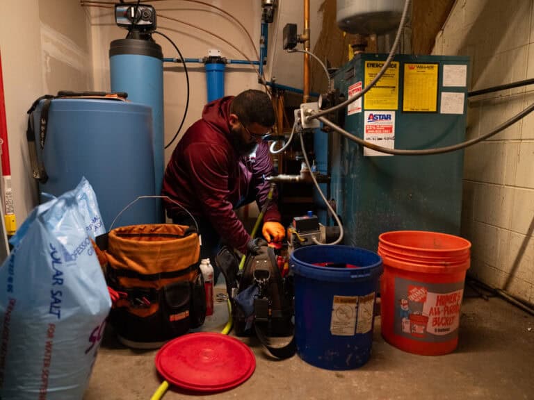 A worker in a maroon hoodie repairs equipment in a utility room, surrounded by tools, blue tanks, orange buckets, and a bag of salt.
