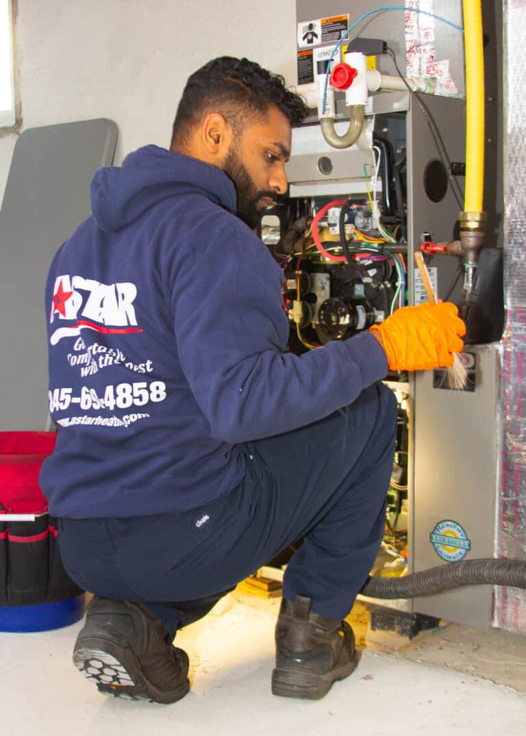A technician wearing a navy hoodie and orange gloves works on a furnace unit, using a screwdriver and inspection brush in a utility room.