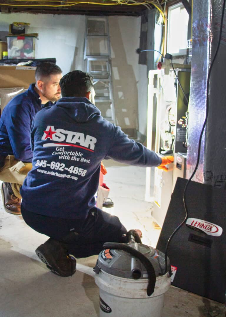 Two technicians inspect and work on a Lennox furnace in a basement, with tools and a vacuum on the floor nearby.