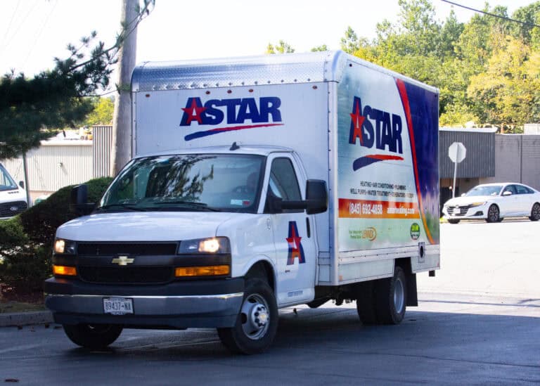 A white Astar delivery truck is parked on a street near a building, with trees and a stop sign in the background.