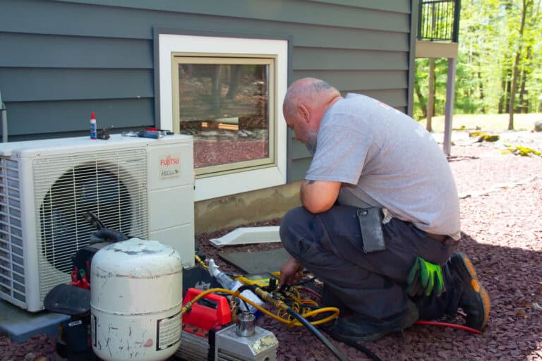 A technician kneels outside a house, servicing an air conditioning unit with various tools and equipment placed nearby.