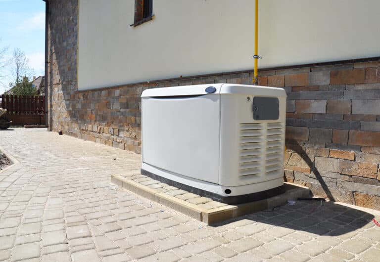 A standby generator is installed on a concrete platform next to a house with stone siding and a paved walkway.
