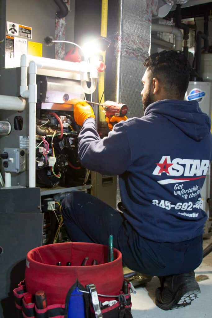 A technician in a blue hoodie uses a power drill to repair HVAC equipment; a red tool bag with various tools is on the floor nearby.