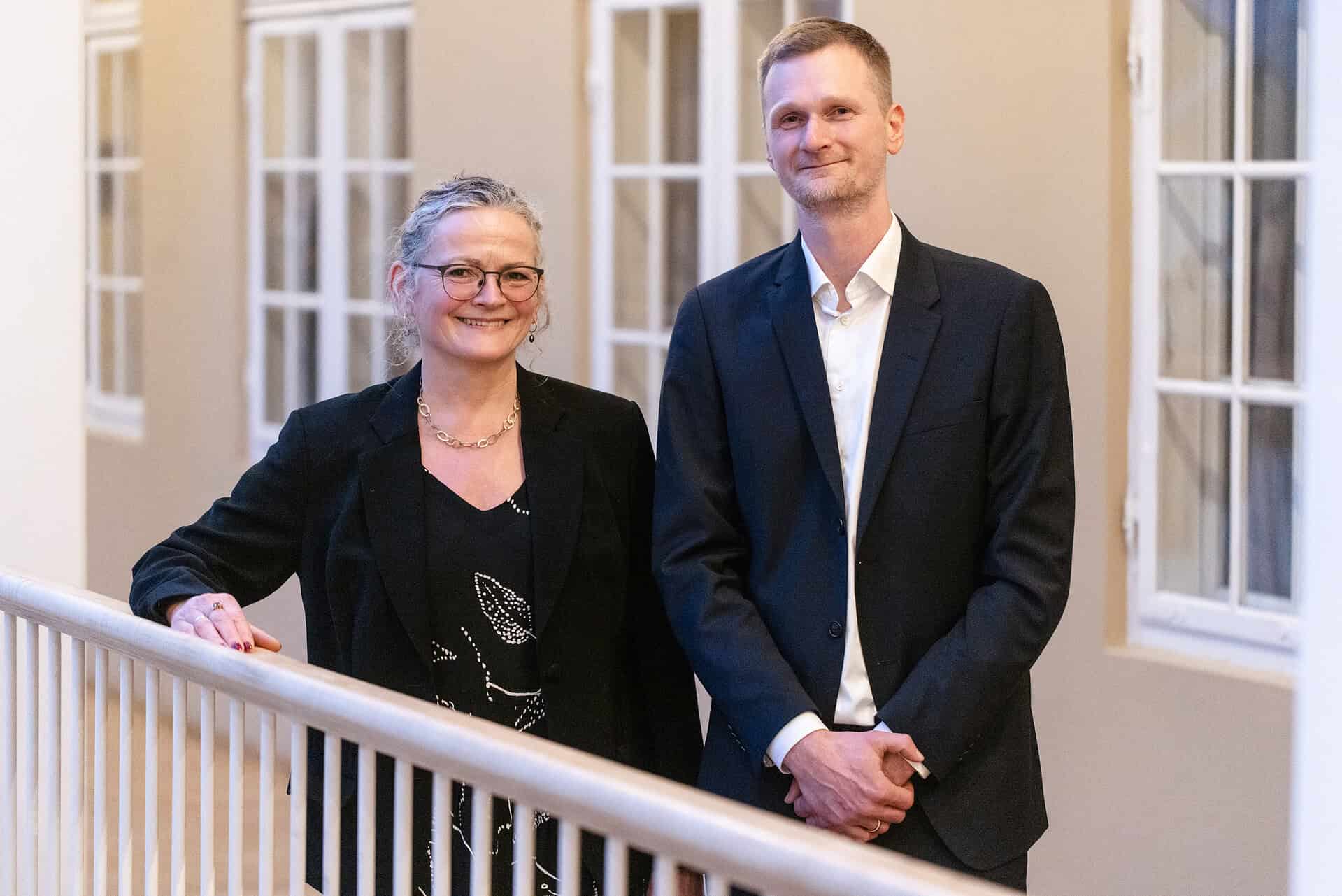 To personer står ved en gelænder i en bygning, smilende og professionelle.