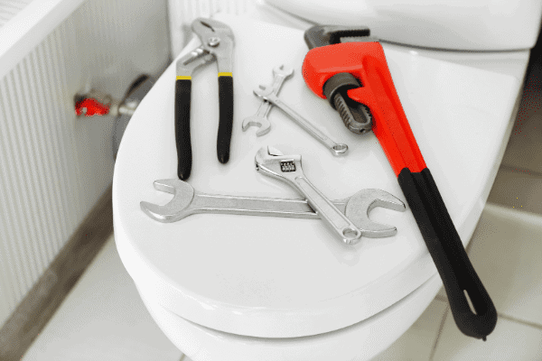 Various plumbing tools, including pliers, wrenches, and a pipe wrench, are placed on a closed toilet lid in a bathroom.
