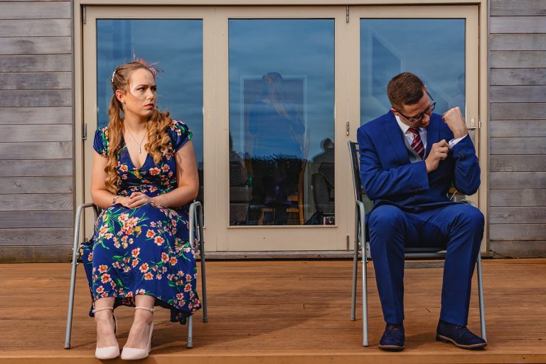 Confused woman in floral dress and man adjusting watch during wedding photo shoot on wooden terrace.