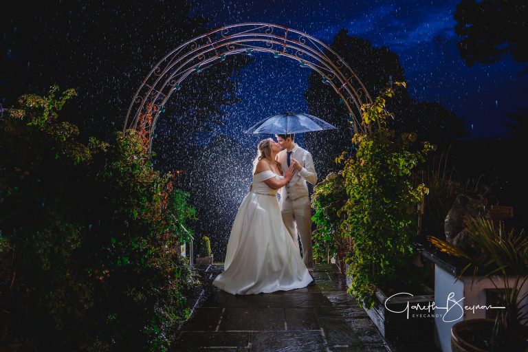 Elegant newlyweds sharing a romantic moment under an umbrella during a rainstorm at night, captured beautifully by Eyecandy Weddings for a memorable wedding photoshoot.