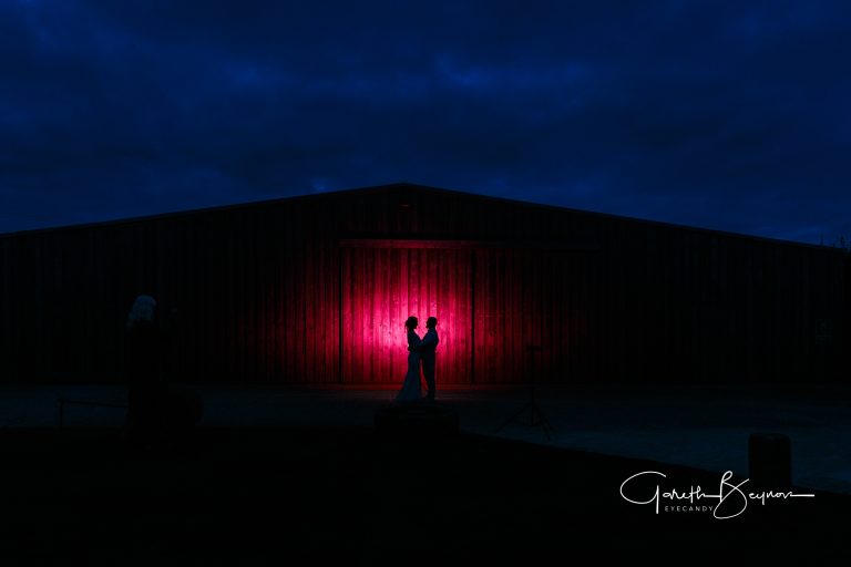 Silhouetted couple sharing a romantic moment against a vibrant pink-lit barn at dusk.