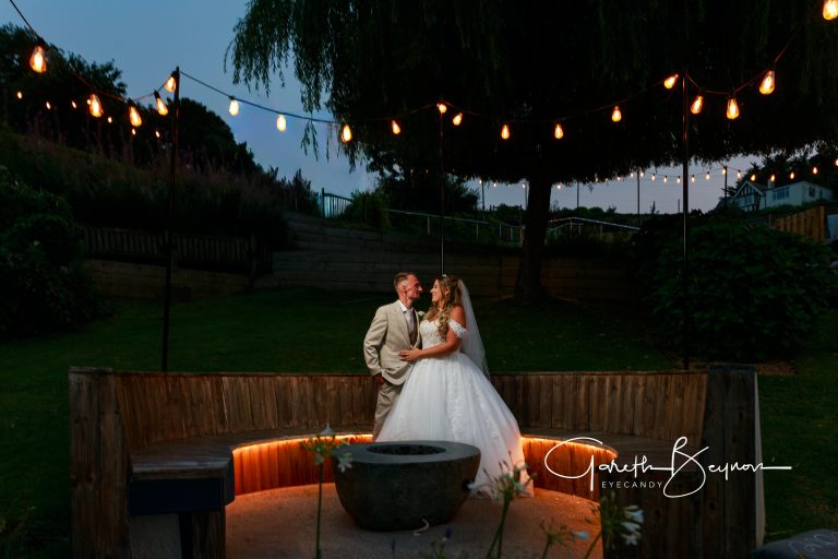Elegant bride and groom sharing a romantic moment during their outdoor wedding at dusk with string lights overhead.