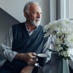 Elderly man looking out the window with a cup of coffee in hand and a vase of white flowers nearby, representing legal support for seniors and elder law services.