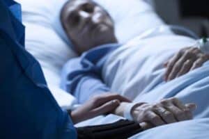 Person lying in hospital bed with oxygen tubes in nose and another person resting their hand on their arm