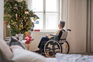 Christmas tree decorated with ornaments and gifts, with an elderly woman in a wheelchair admiring it near a large window. Festive holiday scene emphasizing care and support for seniors during the holiday season.
