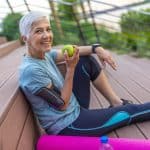 Active senior woman exercising outdoors, holding a green apple, with workout gear and a yoga mat, promoting health and wellness for seniors.