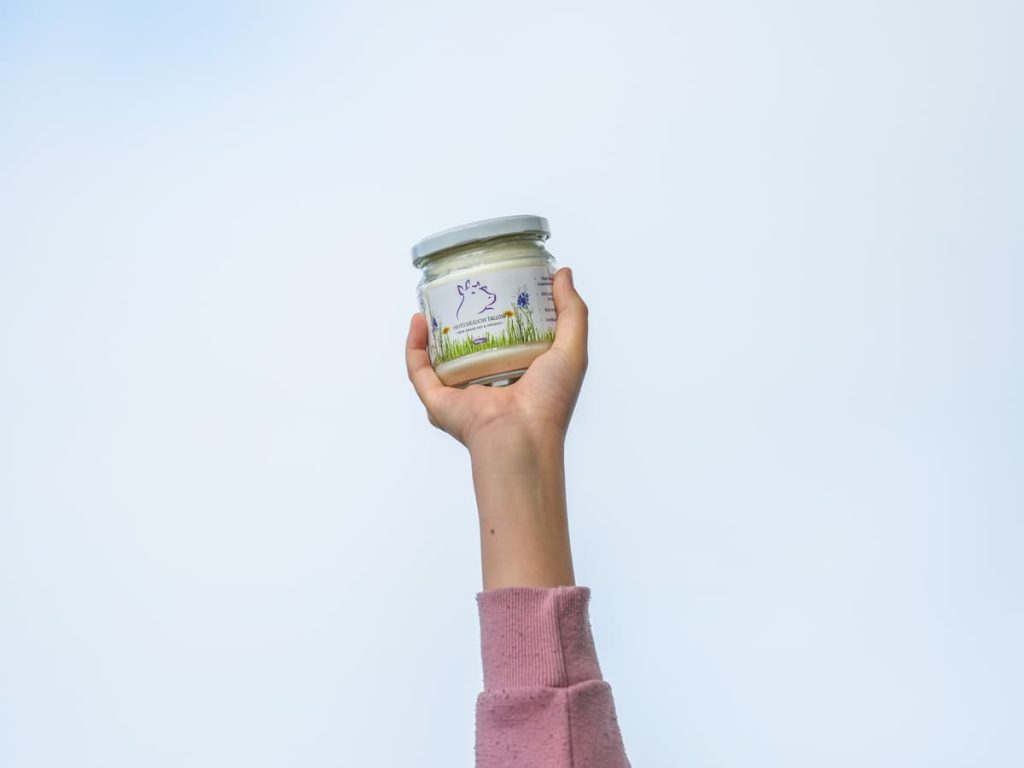 Pure tallow skincare product in a glass jar, held up by a hand against a light blue background.