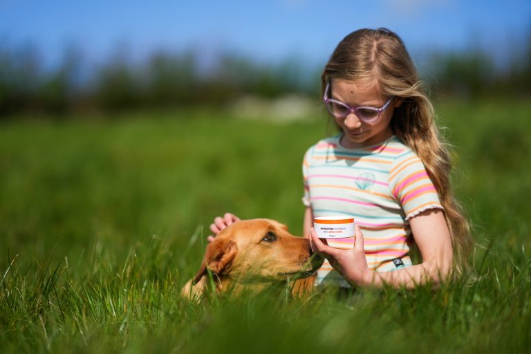 Girl with dog holding organic tallow skincare jar outdoors in a grassy field.