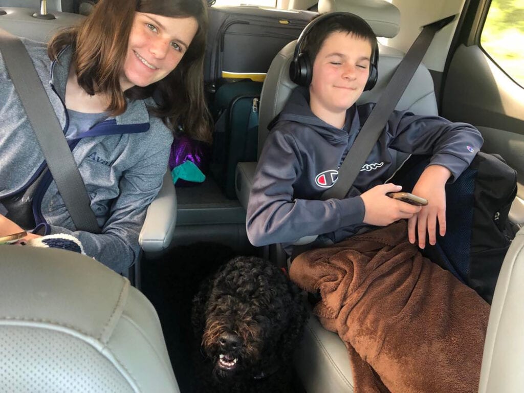 A Stress-Free Family Road Trip Packing List This image shows a boy and a girl sitting in the back seat of a car with a dog. The boy has headphones on and there is luggage behind the kids.
