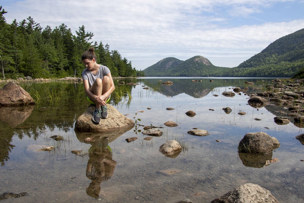 Best Hikes in Acadia National Park + Tips A girl sitting on a large rock in the water at Jordan Pond in Acadia. You can see her reflection in the clear shallow water.