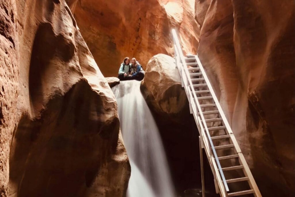 Zion National Park To Horseshoe Bend: A Road Trip Adventure A beautiful waterfall with crystal-clear water cascading down over rocks in a narrow slot canyon. A silver metal staircase leans against the canyon wall near the waterfall, leading to the upper portion of the falls. Sunlight streams in from above, casting a warm glow on the water and surrounding canyon walls.