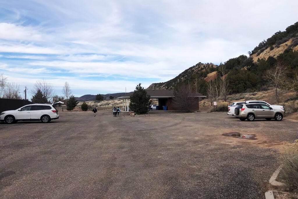 Kanarra Falls: Hike Utah’s Best Slot Canyon A view of the Kanarra Falls parking lot, showing a large paved area with several cars parked and a small building on the right-hand side. There are also picnic tables and restrooms in or by the brick building pictured and a shoe-washing station near the entrance and behind the building.