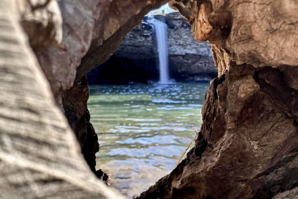 Hiking Desoto Falls? Here’s What You Need to Know View from the base of DeSoto Falls, framed by a natural wooden aperture. The waterfall cascades gracefully, surrounded by lush greenery. The perspective is captured through a unique opening in a wooden structure, adding a natural frame to the scenic beauty.