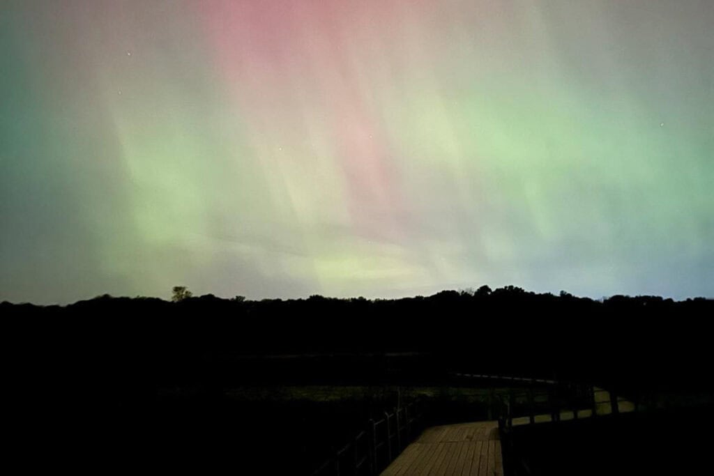 Where to See Northern Lights in Michigan: Upper & Lower Peninsula Green, yellow and pink northern lights shine down on a wooden bridge with trees in the background