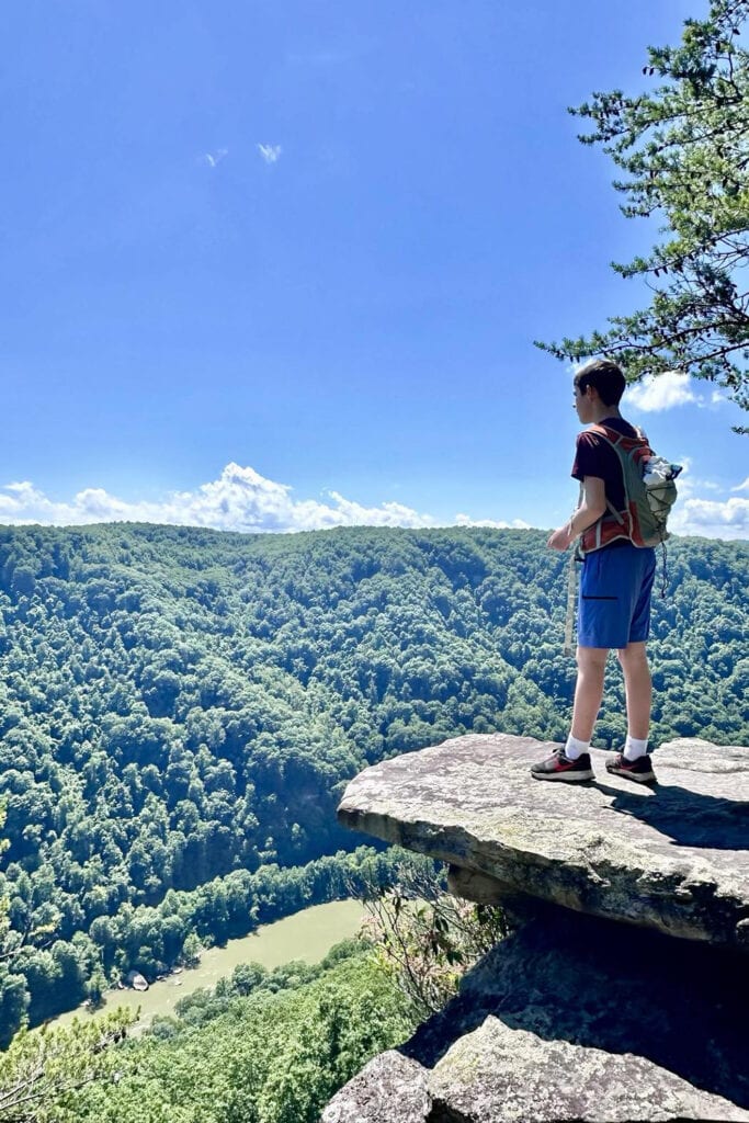 Endless Wall Trail Guide: Trail Tips, Parking, and Scenic Overlooks Boy standing on a rock that over hangs the New River Gorge. He's taking in the view of the New River