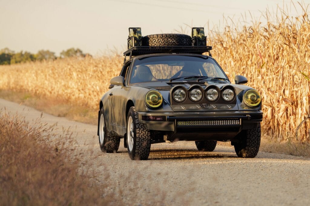 Modified Porsche 964 with off-road tires and roof rack on rural dirt road.