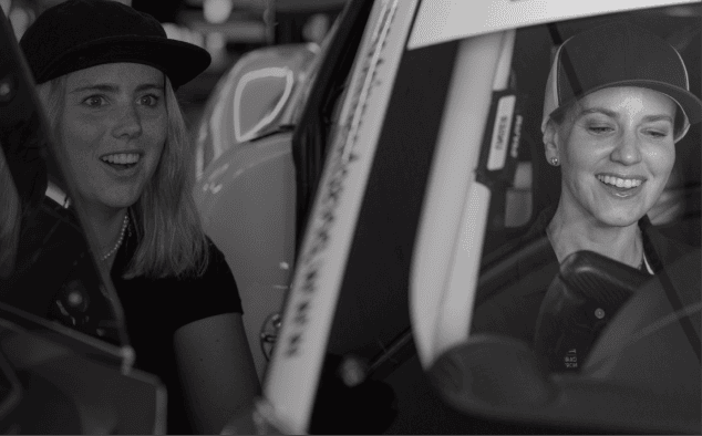 Two women smiling inside a vehicle, one wearing a cap, in black and white.