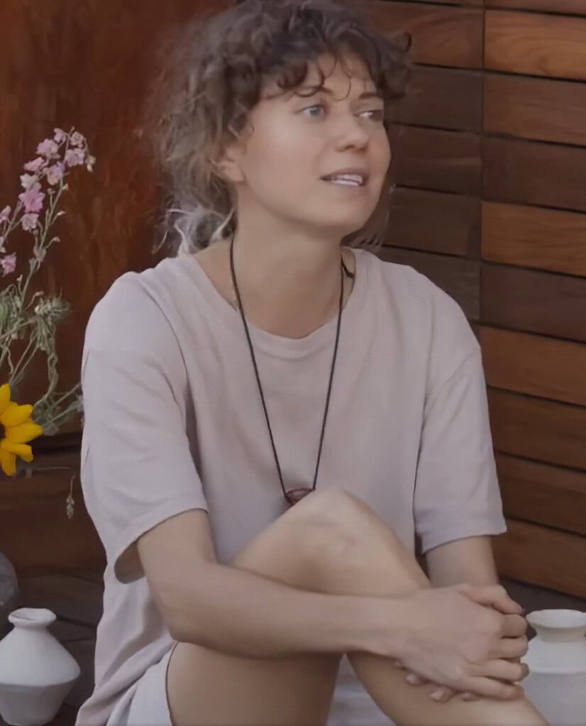 Woman with curly hair sitting outdoors near small ceramic vases and flowers, speaking or listening attentively
