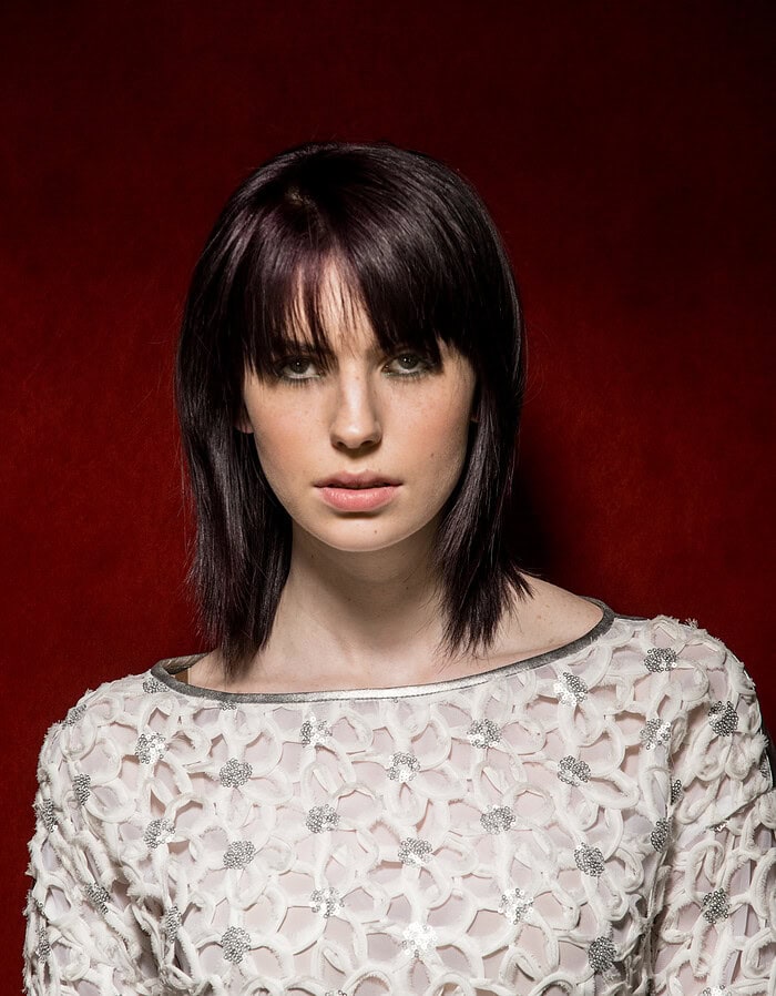 Woman with a sleek dark plum bob haircut with wispy bangs against a deep red background