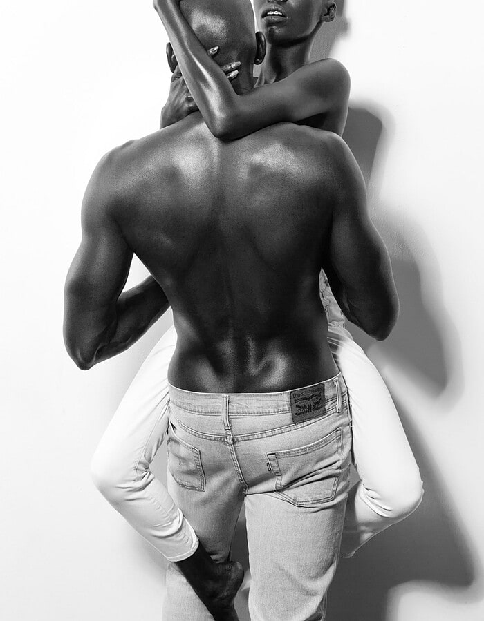 Female model embracing a male model from behind in a black-and-white fashion portrait.