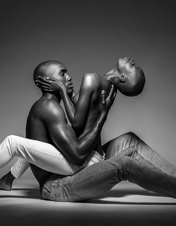 Two models seated in an intertwined pose in a dramatic black-and-white studio photograph.