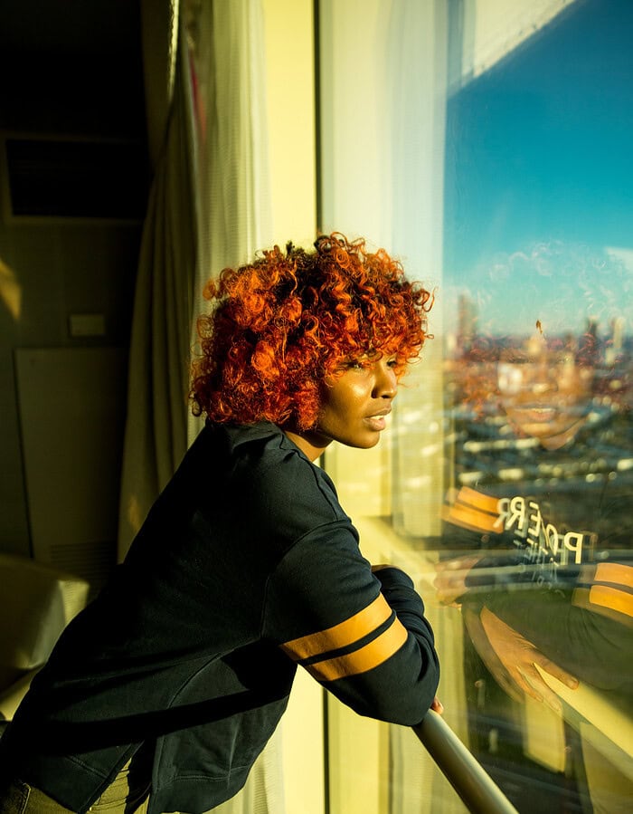 Woman with vibrant red curly hair leaning on a railing, gazing out a large window at a city skyline