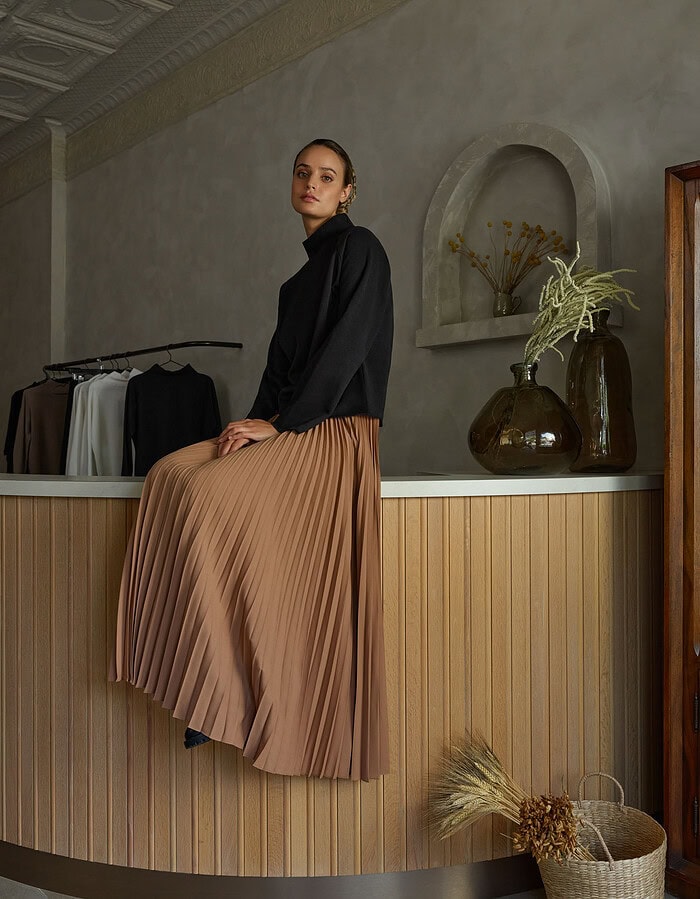 Elegant woman in black top and pleated beige skirt sitting on a wooden counter. photographer NYC