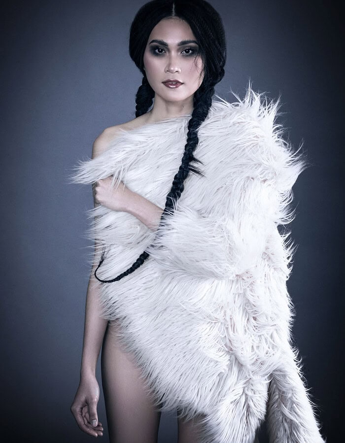 Fashion model wearing a white shaggy fur wrap with long black braids against a dark gray studio background.