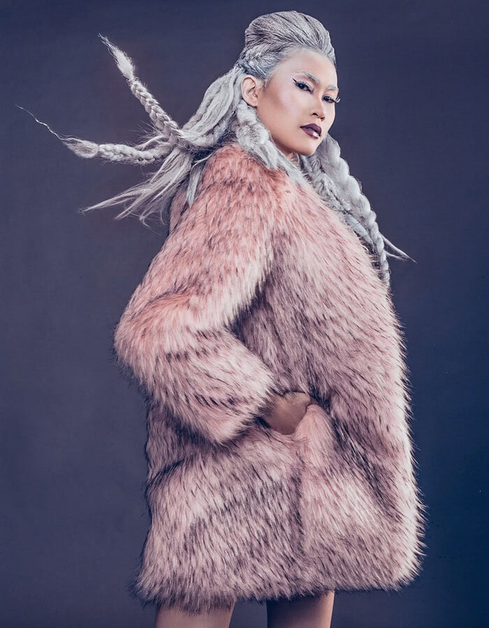 Fashion model in a pink fur coat with silver braids moving through the air against a dark studio background.