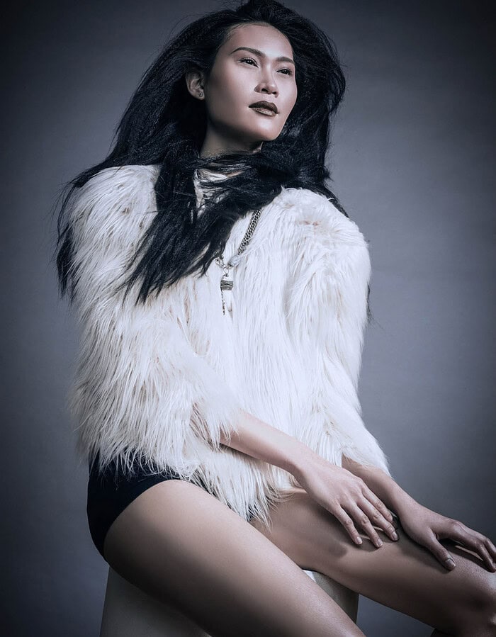 Seated fashion model with long black textured hair wearing a white shaggy fur jacket.