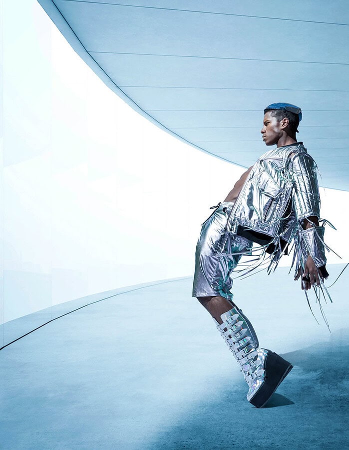 Male fashion model in a metallic silver outfit and platform boots leaning beneath a curved futuristic ceiling.