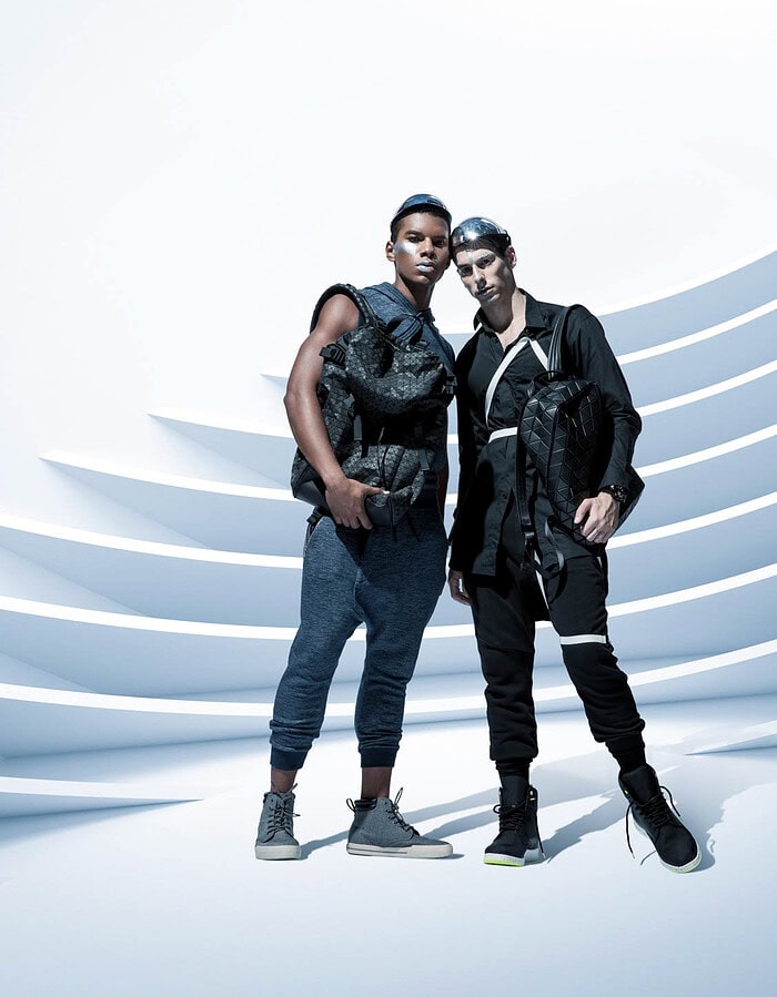 Two male fashion models in black futuristic outfits standing together before curved white architectural forms.