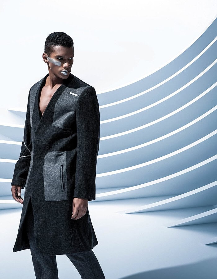 Male fashion model in a long black and gray tailored coat posed against sweeping white architectural curves.