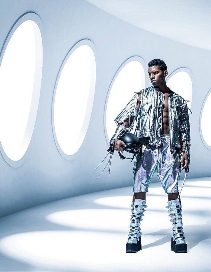 Male fashion model in a metallic silver outfit and platform boots holding a black helmet in a white futuristic corridor.