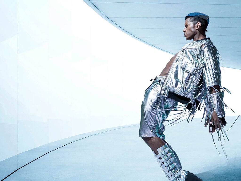 Male fashion model in a silver outfit and platform boots leaning across a cool blue futuristic set.