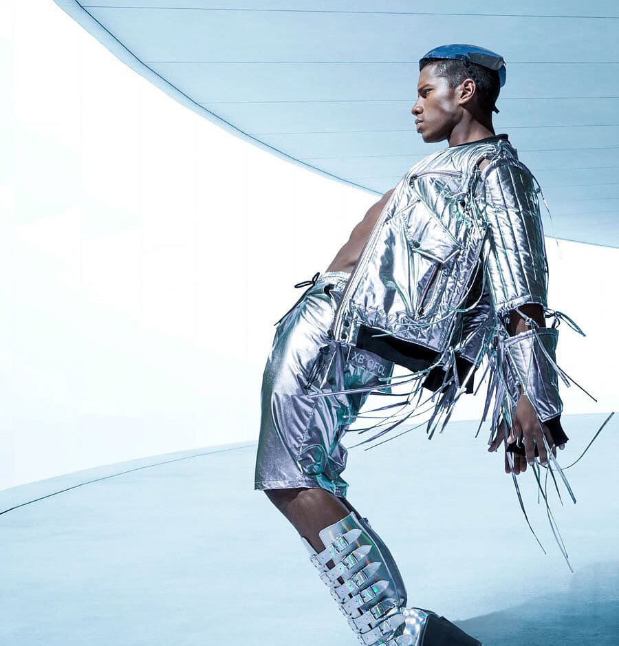 Male fashion model in a silver outfit and platform boots leaning across a cool blue futuristic set.