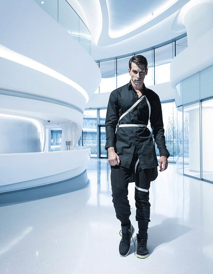 Male fashion model in a black outfit with white straps standing in a bright curved modern interior.