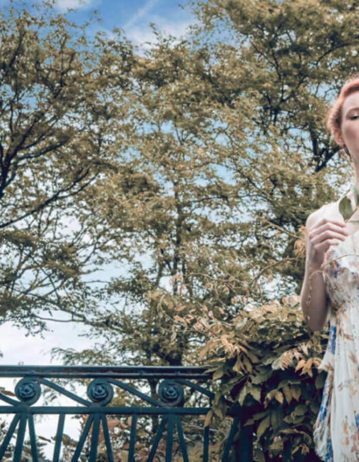 Red-haired fashion model wearing a flowing floral dress beside an iron railing in a tree-filled garden.