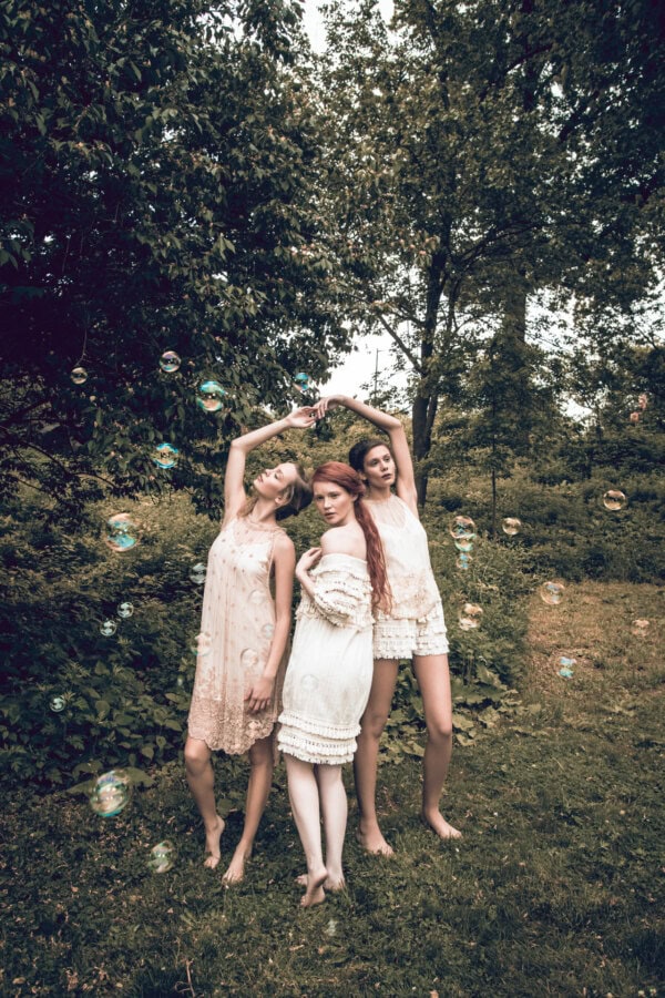 Three fashion models in pale dresses posing barefoot in a wooded garden surrounded by floating bubbles.