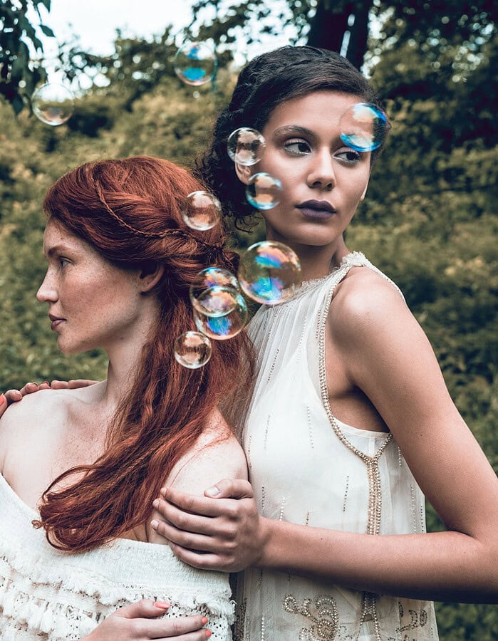 Three fashion models in pale dresses posed closely together as bubbles float through a green garden.