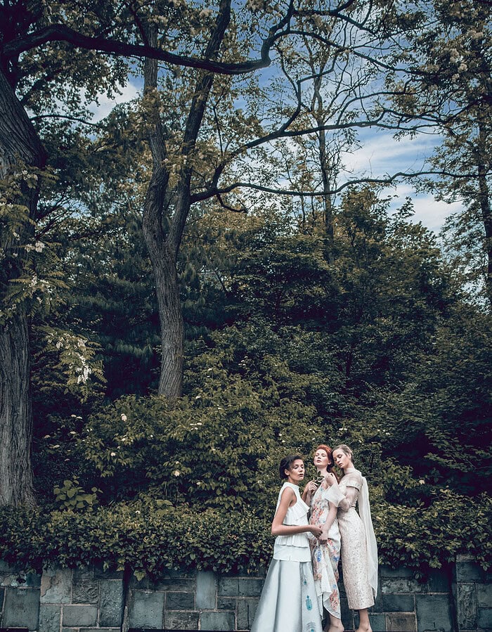 Three barefoot fashion models in pale dresses standing together on a park bench beneath tall trees.