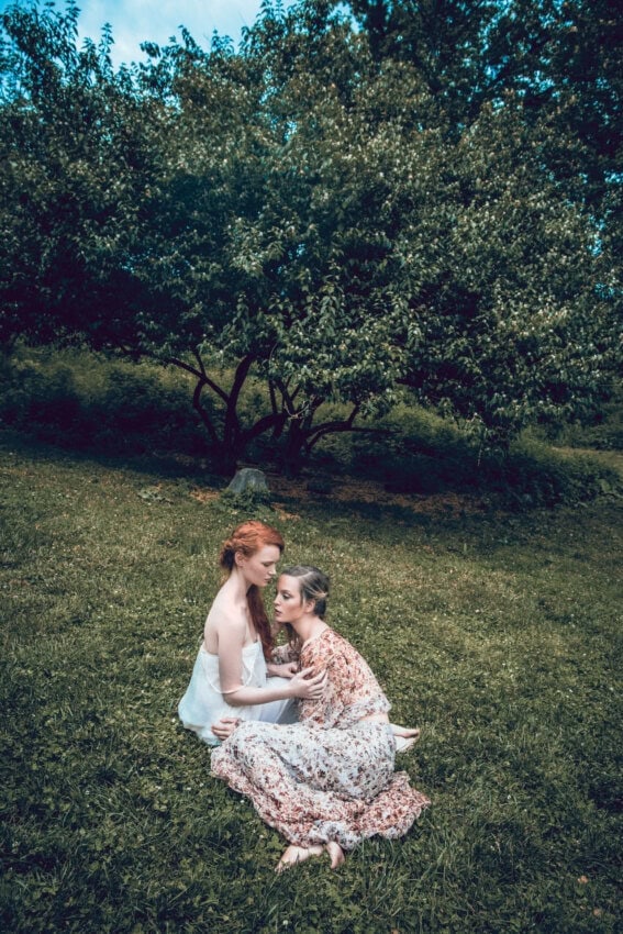 Two fashion models in flowing pale floral dresses seated together on the grass in a wooded meadow.