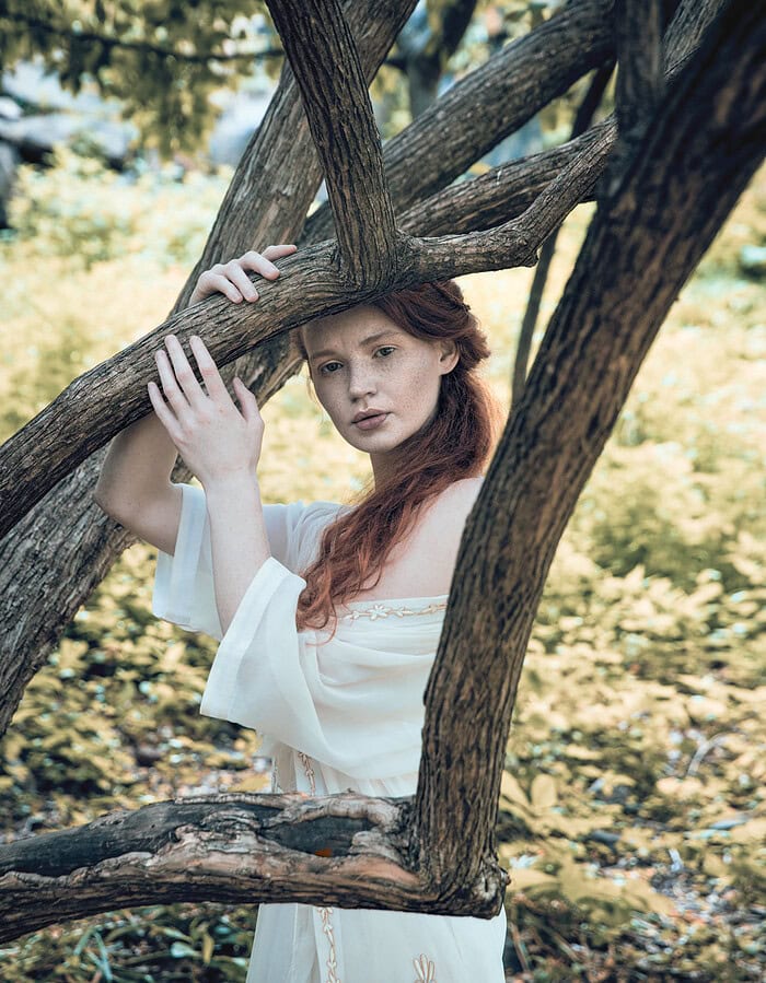 Red-haired fashion model in a white embroidered dress framed by twisting tree branches.